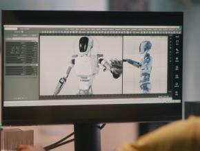 A person sits at a computer that is displaying a robot on the screen